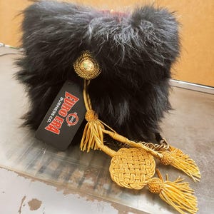 Può includere: Un cappello di pelliccia nera con nappe dorate e un bottone decorativo. Il cappello ha un'etichetta con la scritta "EURO ERA BUSINESS CO.". Le nappe sono attaccate al cappello con una catena d'oro. Il cappello è probabilmente di stile militare o cerimoniale.