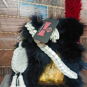 Può includere: Un cappello di pelliccia nero con base color oro e pennacchio rosso. Il cappello presenta un dettaglio in corda bianca e un'etichetta con la scritta "EURO ERA BUSINESS CO."
