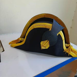 May include: A black bicorne hat with gold trim and tassels. The hat features a decorative black emblem on the side and gold accents along the brim. The hat is likely a historical or costume piece.
