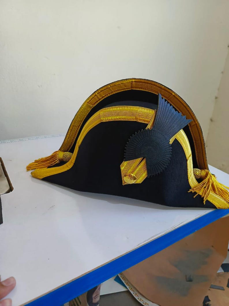 May include: A black bicorne hat with gold trim and tassels. The hat features a decorative black emblem on the side and gold accents along the brim. The hat is likely a historical or costume piece.