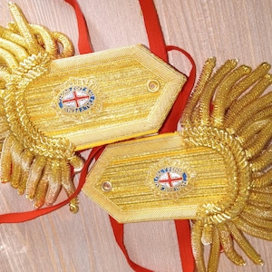 May include: Two gold-colored military shoulder boards with intricate detailing. Each board features a central emblem with a red cross and blue accents, surrounded by text. Red ribbons are attached. The boards are on a light-colored surface.
