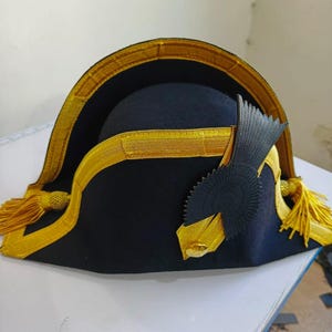 May include: A black bicorne hat with gold trim and tassels. The hat has a decorative black emblem on the side. The brim is trimmed with gold fabric. The hat is likely a historical or costume piece.