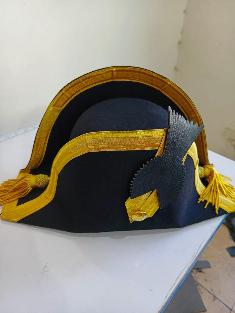 May include: A black bicorne hat with gold trim and tassels. The hat has a decorative black emblem on the side. The brim is trimmed with gold fabric. The hat is likely a historical or costume piece.
