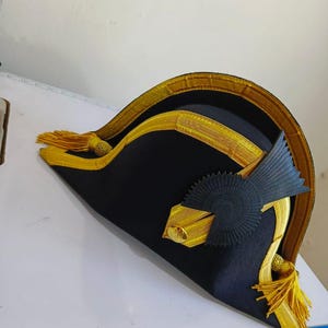 May include: A black bicorne hat with gold trim and tassels. The hat features a decorative gold emblem and a black rosette. The hat is likely a historical or costume piece.
