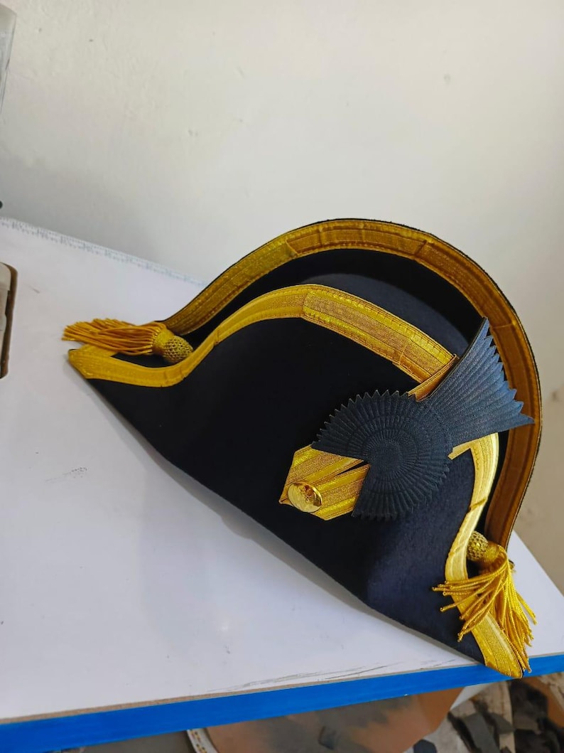 May include: A black bicorne hat with gold trim and tassels. The hat features a decorative gold emblem and a black rosette. The hat is likely a historical or costume piece.