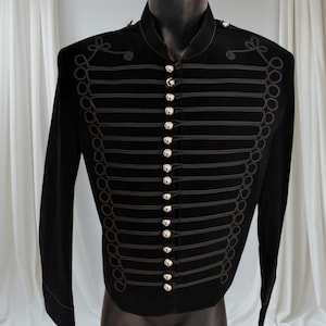Peut inclure: Veste noire de style militaire avec un col montant et des manches longues. La veste présente des rangées de garnitures horizontales noires et des boutons argentés sur le devant. Des garnitures noires décoratives ornent les épaules et les côtés.