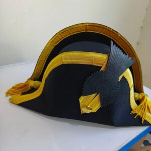 May include: A black bicorne hat with gold trim and tassels. The hat has a gold band around the brim and a decorative black and gold emblem on the side. The hat is a historical style.