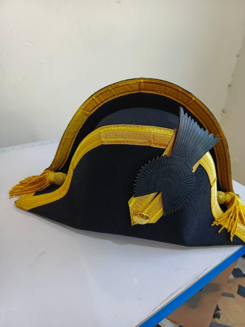 May include: A black bicorne hat with gold trim and tassels. The hat has a gold band around the brim and a decorative black and gold emblem on the side. The hat is a historical style.