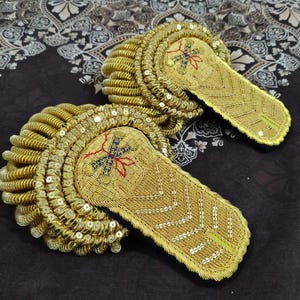 Op de afbeelding: Twee goudkleurige epauletten met ingewikkelde borduursels en pailletten details. Elke epaulet heeft een centraal ontwerp met rode, blauwe en gouden draden. De randen zijn zorgvuldig afgewerkt met opgerolde gouden draad.