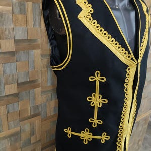 May include: Black vest with gold trim and decorative floral embroidery. The vest has a V-neck and is sleeveless. The gold trim outlines the edges and the floral embroidery is on the front. The vest is displayed on a mannequin.