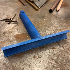Puede incluir: Una herramienta azul en forma de T con una superficie texturizada y un mango cil&iacute;ndrico unido a una barra perpendicular. El objeto presenta un peque&ntilde;o orificio en la parte superior del mango y un borde c&oacute;ncavo a lo largo de la barra horizontal.