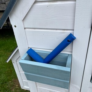 Puede incluir: Una jardinera rectangular de madera azul claro montada en una pared blanca de paneles horizontales. Dentro de la jardinera hay una herramienta de pl&aacute;stico azul brillante con un mango largo y un orificio circular en la parte superior, inclinada.