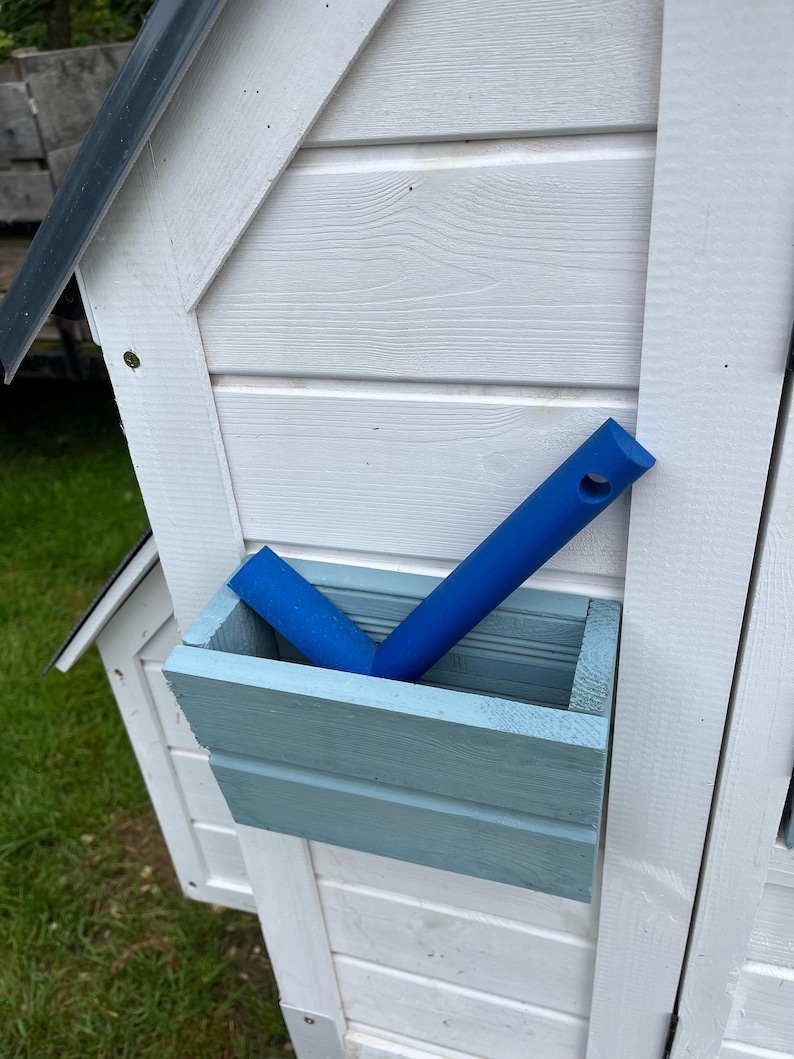 Puede incluir: Una jardinera rectangular de madera azul claro montada en una pared blanca de paneles horizontales. Dentro de la jardinera hay una herramienta de pl&aacute;stico azul brillante con un mango largo y un orificio circular en la parte superior, inclinada.