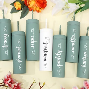 May include: A collection of personalized tumblers in shades of green and white, each with a straw and a name in elegant script. The central white tumbler is labeled "Bride". The tumblers are arranged amidst colorful flowers.