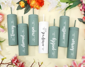 Op de afbeelding: Een verzameling gepersonaliseerde bekers in groen en wit, elk met een rietje en een naam in elegant schrift. De centrale witte beker is voorzien van het opschrift "Bride". De bekers zijn gerangschikt tussen kleurrijke bloemen.