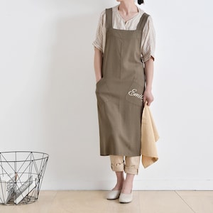 May include: A long, olive-green apron with large front pockets and a white embroidered name. The apron has wide shoulder straps and is worn over a striped shirt and tan pants. A folded tan towel is held in one hand.