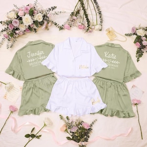 Puede incluir: Dos conjuntos de pijama verde salvia con texto blanco Jennifer y Katie, y un conjunto blanco con texto dorado Natalie y Bride. Cada conjunto incluye una camisa abotonada con mangas con volantes y pantalones cortos a juego con volantes.