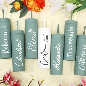 May include: A collection of personalized tumblers in shades of green and white, arranged with floral accents. The tumblers feature names like "Rebecca," "Charlotte," and "Catherine," with one labeled "Bride." Each tumbler has a straw.
