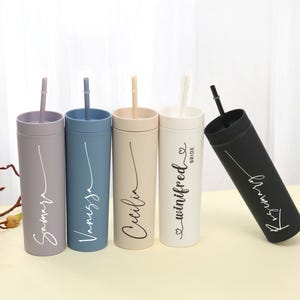 May include: A collection of personalized tumblers in various colors, including gray, blue, beige, white, and black. Each tumbler has a matching straw and features a name in a cursive font. One tumbler is labeled "Bride".