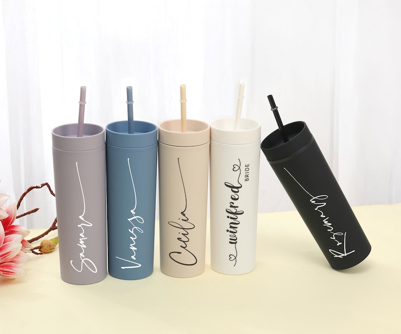 May include: A collection of personalized tumblers in various colors, including gray, blue, beige, white, and black. Each tumbler has a matching straw and features a name in a cursive font. One tumbler is labeled "Bride".