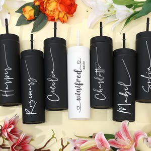 May include: A collection of personalized black and white tumblers with straws, arranged amidst flowers. Each tumbler is inscribed with a name in elegant script, with one tumbler reading "Bride". The tumblers are cylindrical and appear to be made of metal.