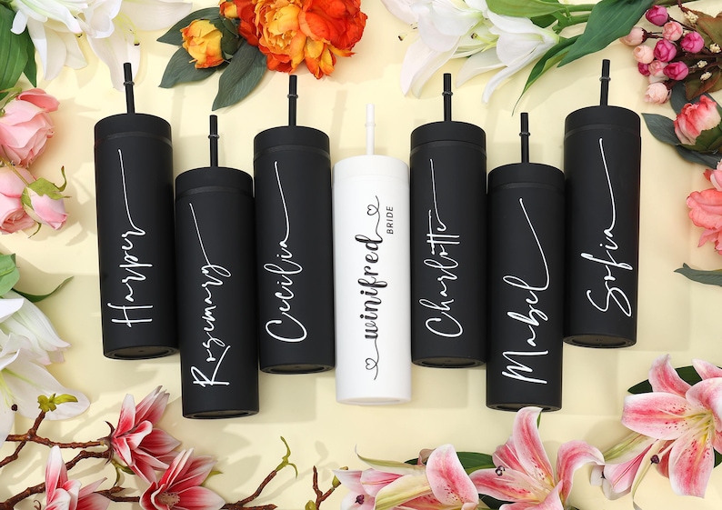 May include: A collection of personalized black and white tumblers with straws, arranged amidst flowers. Each tumbler is inscribed with a name in elegant script, with one tumbler reading "Bride". The tumblers are cylindrical and appear to be made of metal.