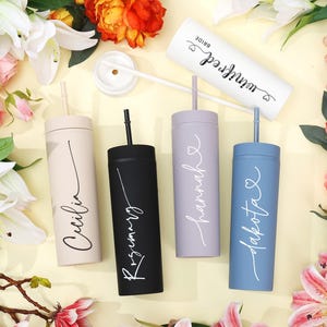 May include: A collection of personalized tumblers in various colors, including beige, black, lavender, and blue, each with a name in elegant script. One white tumbler reads "Bride" and "Winifred". The tumblers have straws and are surrounded by flowers.