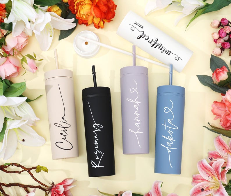 May include: A collection of personalized tumblers in various colors, including beige, black, lavender, and blue, each with a name in elegant script. One white tumbler reads "Bride" and "Winifred". The tumblers have straws and are surrounded by flowers.