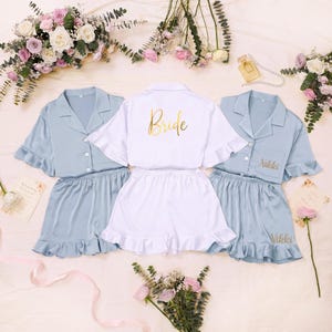 Puede incluir: Dos conjuntos de camisas de pijama abotonadas y pantalones cortos de color azul claro con dobladillos con volantes, y un conjunto de camisa de pijama abotonada y pantalones cortos blancos con volantes. La camisa blanca dice Bride en oro y las azules Nikki.