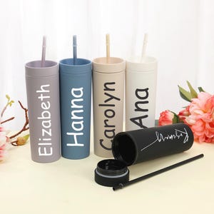 May include: A collection of personalized tumblers in various colors, including lavender, blue, beige, and white. Each tumbler has a name printed on it in a contrasting color. One black tumbler is open, with a straw and lid visible.