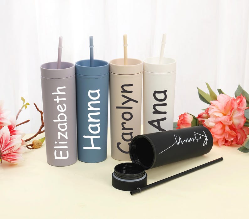 May include: A collection of personalized tumblers in various colors, including lavender, blue, beige, and white. Each tumbler has a name printed on it in a contrasting color. One black tumbler is open, with a straw and lid visible.