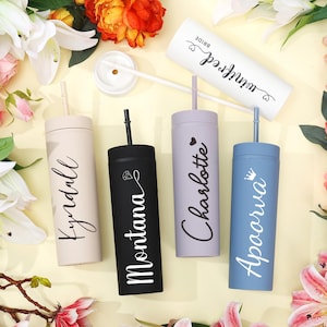 May include: A collection of personalized tumblers in various colors, including beige, black, lavender, and blue. Each tumbler features a name in elegant script. The tumblers are arranged with flowers, suggesting a celebratory theme.