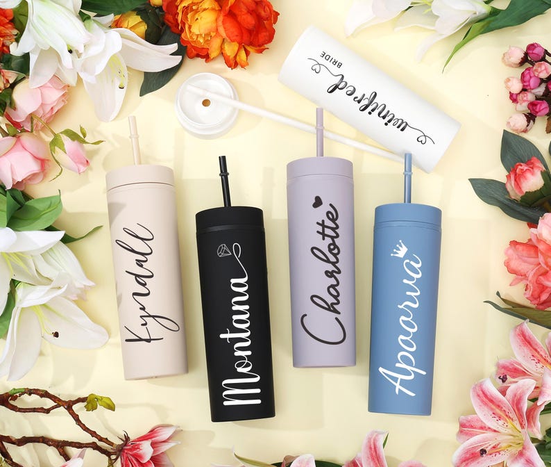 May include: A collection of personalized tumblers in various colors, including beige, black, lavender, and blue. Each tumbler features a name in elegant script. The tumblers are arranged with flowers, suggesting a celebratory theme.