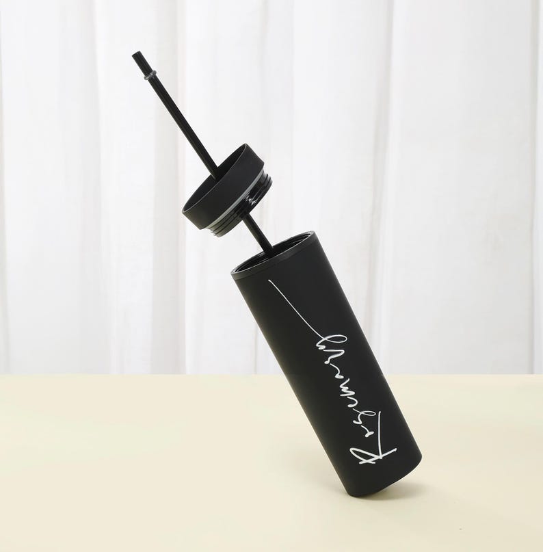 May include: A black tumbler with a matching lid and straw. The tumbler has white cursive text on the side. The lid is off, and the straw is angled upwards. The tumbler is cylindrical and appears to be made of metal.