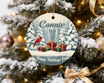 May include: Round Christmas ornament with a winter scene featuring a red cardinal, snow-covered trees, and wrapped gifts. The ornament has the name "Connie" and the year "2025" printed on it, with the text "Happy Holidays from".