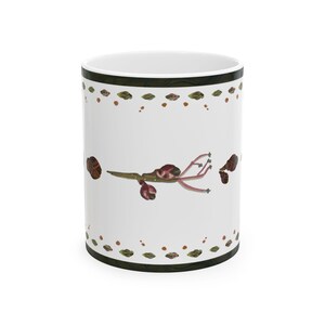 May include: A white ceramic mug featuring a central illustration of a brown twig with small reddish-brown buds and delicate stems. The top and bottom rims are decorated with a repeating pattern of tiny green leaves and brown dots on a dark border.
