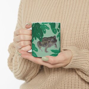May include: A white ceramic mug featuring a printed illustration of a brown toad with dark spots positioned among vibrant green leaves. The design covers the exterior surface of the mug, showcasing a detailed nature-inspired graphic.