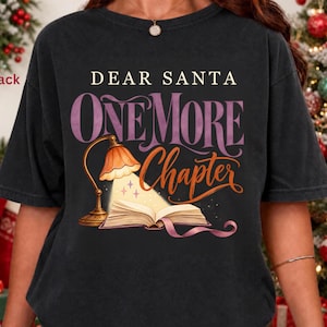Może przedstawiać: Czarny t-shirt z krótkim rękawem z napisem DEAR SANTA ONE MORE CHAPTER w kolorze białym, fioletowym i pomarańczowym. Grafika przedstawia zapaloną lampkę biurkową oświetlającą otwartą książkę z małymi akcentami w kształcie gwiazdek.