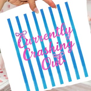 May include: White square pillow with blue vertical stripes and the text "Currently Crashing Out" in pink script. The text includes decorative bow accents. The pillow is held by a hand.