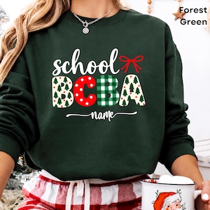 Op de afbeelding: Een bosgroene sweater met ronde hals en de witte tekst school en BCBA in een decoratief lettertype. De letters zijn gevuld met patronen van kerstbomen, rode stippen en groen ruitpatroon. Een rode strik en het woord name staan eronder.
