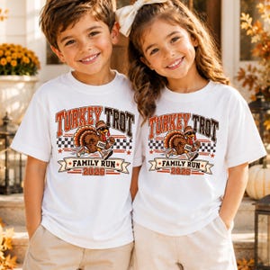May include: Two white short-sleeved t-shirts featuring a graphic of a running turkey wearing sunglasses and sneakers. The text reads Turkey Trot Family Run 2026. The design includes a checkered pattern, stars, and orange and brown lettering.