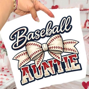 Könnte beinhalten: Weißes quadratisches Kissen mit Baseball-Motiv. Das Design zeigt das Wort "Baseball" in marineblauer Schrift, eine schleife in Form eines Baseballs und das Wort "AUNTIE" in roten und beigen Blockbuchstaben. Das Kissen wird von einer Person gehalten.