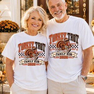 May include: Two white t-shirts featuring a graphic of a running turkey wearing sunglasses and sneakers. The text reads Turkey Trot Family Run 2026. The design includes a checkered pattern, orange and brown lettering, and star accents on the shirts.