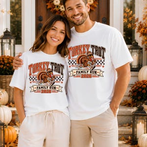 May include: Two white t-shirts featuring a graphic print of a turkey wearing sunglasses and sneakers. The text reads Turkey Trot Family Run 2026. The design includes a checkered pattern, stars, and orange and brown lettering on the front.