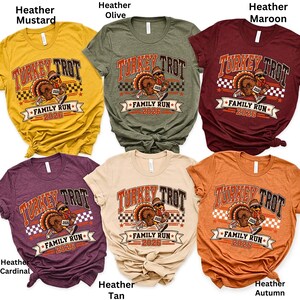 May include: Six short-sleeved t-shirts in heather mustard, heather olive, heather maroon, heather cardinal, heather tan, and heather autumn. Each shirt features a graphic of a running turkey wearing a hat and sneakers, with the text Turkey Trot Family Run 2026.