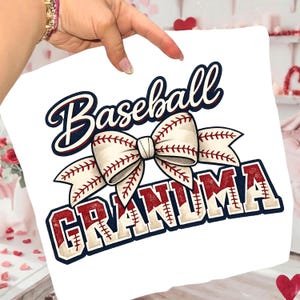 May include: White cushion featuring the text "Baseball Grandma" in a script font, with a baseball-stitched bow. The text is in red, white, and blue. A baseball-themed gift.