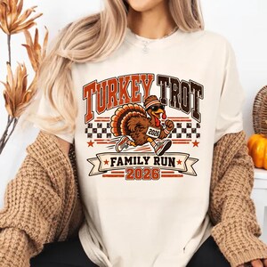 May include: A cream-colored t-shirt featuring a graphic of a running turkey wearing sunglasses, a headband, and sneakers. The text reads "TURKEY TROT", "FAMILY RUN", and "2026". The design includes a checkered pattern and orange star accents.