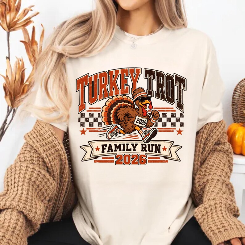 May include: A cream-colored t-shirt featuring a graphic of a running turkey wearing sunglasses, a headband, and sneakers. The text reads "TURKEY TROT", "FAMILY RUN", and "2026". The design includes a checkered pattern and orange star accents.