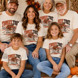 May include: A group of people wearing matching cream-colored t-shirts featuring a graphic of a turkey wearing a hat and running shoes. The text reads Turkey Trot Family Run 2026, accompanied by a checkered pattern and red and orange accents.