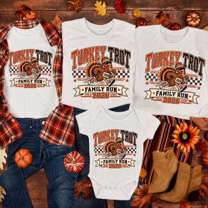 May include: Three white t-shirts and one white baby bodysuit featuring a graphic of a running turkey wearing a hat and sneakers. The text reads Turkey Trot Family Run 2026. The design includes a checkered pattern and orange and brown accents.